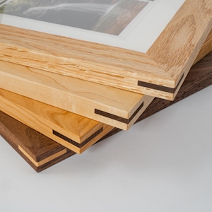 May include: Close-up of four wooden picture frames stacked on top of each other. The frames are made of different types of wood, including oak and walnut. The frames have a simple, classic design and are perfect for displaying photos or artwork.