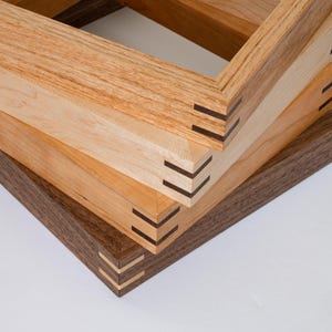 May include: Three wooden picture frames stacked on top of each other. The top frame is light brown, the middle frame is a medium brown, and the bottom frame is a dark brown. All three frames have a mitered joint with a black inlay.