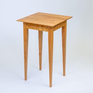 May include: A small, square-topped wooden table with four slender, tapered legs. The table is made of light-colored wood, possibly maple or birch, and has a natural finish. The legs are evenly spaced and provide a stable base.