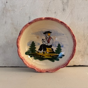 May include: A small, round ceramic bowl with a scalloped edge. The bowl features a hand-painted scene of a person in traditional attire smoking a pipe, seated on a rock. The rim is red, and the interior has a cream-colored background.