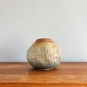May include: A handcrafted ceramic vase with a rounded shape and textured surface. The vase features a blend of earthy tones, including brown, beige, and green. The exterior has vertical and horizontal carved lines. The vase sits on a wooden surface.