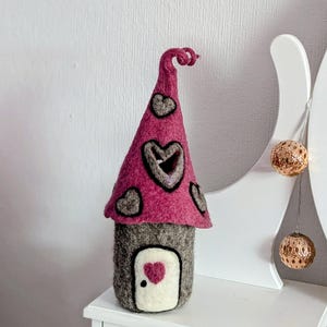 May include: A handcrafted felt house with a pink roof decorated with grey heart shapes. The house has a grey body with a white door featuring a pink heart. The roof has a curled top.