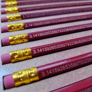 Op de afbeelding: Een close-up van een stapel rode potloden met gouden punten. Elk potlood heeft het getal 3.1415926535897932384626433832795028841971693993751058209749445923078164062862089986280348253421170679 erop gedrukt.