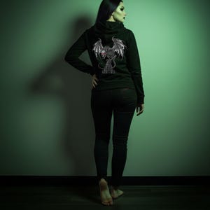 以下が含まれることがあります： 背中にドラゴンのグラフィックが入った黒いジップアップパーカー。ドラゴンは灰色の翼と赤いドクロが特徴です。画像内の人物は黒いジーンズを着用しています。背景は緑色の壁です。