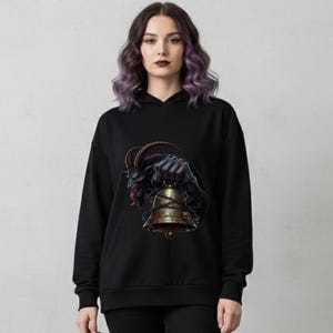 May include: Black hoodie featuring a detailed graphic of a horned demon holding a golden bell. The artwork has a dark, gothic theme with intricate details and a contrasting color palette. The hoodie is a comfortable, casual garment.