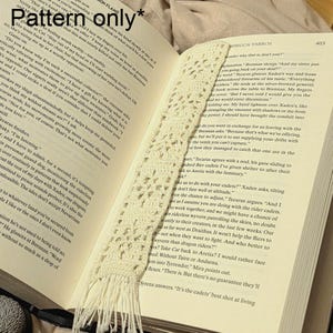 Può includere: Segnalibro all'uncinetto color crema a trama aperta con fondo sfrangiato, appoggiato sulle pagine di un libro. Il delicato motivo e la trama del segnalibro sono visibili contro il testo stampato.