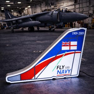 Può includere: Un'insegna decorativa illuminata a forma di deriva di aereo. L'insegna presenta la Union Jack e un design rosso e bianco, con la scritta "FLY 100 NAVY" e gli anni "1909-2009". Un aereo grigio è sullo sfondo.