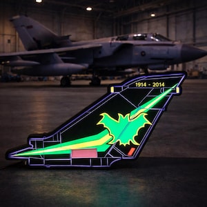 Puede incluir: Un estabilizador de cola de avión negro y verde iluminado con el texto "1914 - 2014". La aleta presenta un emblema verde con forma de murciélago y un diseño amarillo y verde. Un avión es visible en el fondo.