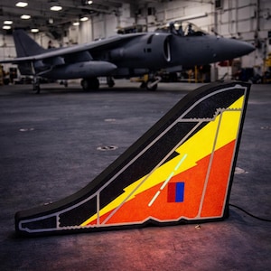 Könnte beinhalten: Ein schwarzes, beleuchtetes, stilisiertes Flugzeugseitenleitwerk mit gelben, orangefarbenen und roten Akzenten. Ein blaues und rotes Rechteck befindet sich auf dem Seitenleitwerk. Im Hintergrund ist ein graues Flugzeug zu sehen.