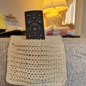 Op de afbeelding: Een beige gehaakte afstandsbedieninghouder, met een zwarte afstandsbediening erin, die op een grijze bank ligt.