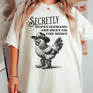 May include: A white t-shirt with a black and white illustration of a chicken wearing a cowboy hat and boots. The text "Secretly Hopes Humans Are Next On The Menu" is printed on the shirt.