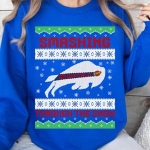 May include: A royal blue sweatshirt with a festive design. The design includes the words "SMASHING THROUGH THE SNOW" in red, a white buffalo with a football, and snowflake accents. The phrase "GO BUFFALO" is also visible.