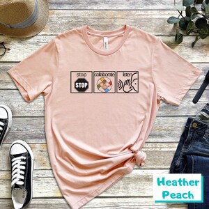May include: A pink t-shirt with a graphic of three icons. The first icon is a stop sign with the word "STOP" inside. The second icon is a circle with five hands of different skin tones holding each other. The third icon is a hand cupping an ear with the word "listen" above it. The text "collaborate" is above the second icon. The text "stop" is above the first icon. The text "listen" is above the third icon. The t-shirt is labeled "Heather Peach".