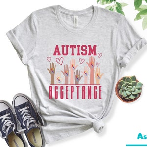May include: A gray t-shirt with the text "AUTISM ACCEPTANCE" in red letters. The design features multiple hands with the infinity symbol in different colors.