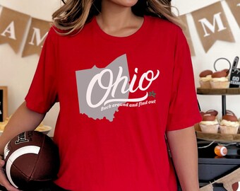 May include: A red t-shirt featuring the state of Ohio in gray with the word "Ohio" in white script. Below the state outline, the text reads "Buck around and find out". The shirt is short-sleeved.