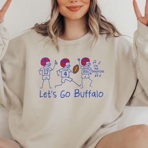 May include: Beige sweatshirt with a blue graphic of three football players and the text "Let's Go Buffalo." The players wear red helmets and are numbered 17, 4, and 86. Additional text includes "I'm Mr. Brightside" and musical notes.
