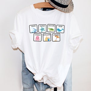K&ouml;nnte beinhalten: Wei&szlig;es T-Shirt mit einer Grafik von bunten Strand-Icons in Quadraten. Die Icons beinhalten Wellen, einen Seestern, eine Meeresschildkr&ouml;te, einen Wal, einen Oktopus, eine Sandburg und einen Regenschirm. Die W&ouml;rter "Wellen", "Seestern", "Meeresschildkr&ouml;te", "Wal", "Oktopus", "Sandburg" und "Regenschirm" sind unter jedem Icon gedruckt.