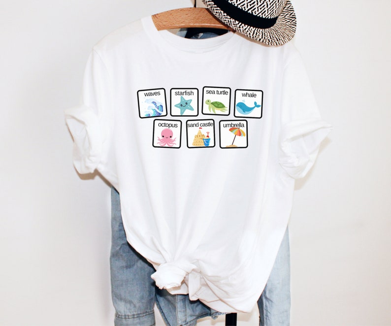 K&ouml;nnte beinhalten: Wei&szlig;es T-Shirt mit einer Grafik von bunten Strand-Icons in Quadraten. Die Icons beinhalten Wellen, einen Seestern, eine Meeresschildkr&ouml;te, einen Wal, einen Oktopus, eine Sandburg und einen Regenschirm. Die W&ouml;rter "Wellen", "Seestern", "Meeresschildkr&ouml;te", "Wal", "Oktopus", "Sandburg" und "Regenschirm" sind unter jedem Icon gedruckt.