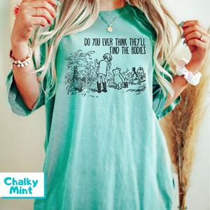 May include: A teal blue t-shirt featuring a black and white illustration of Winnie the Pooh and his friends. The text on the shirt reads "Do you ever think they'll find the bodies?"