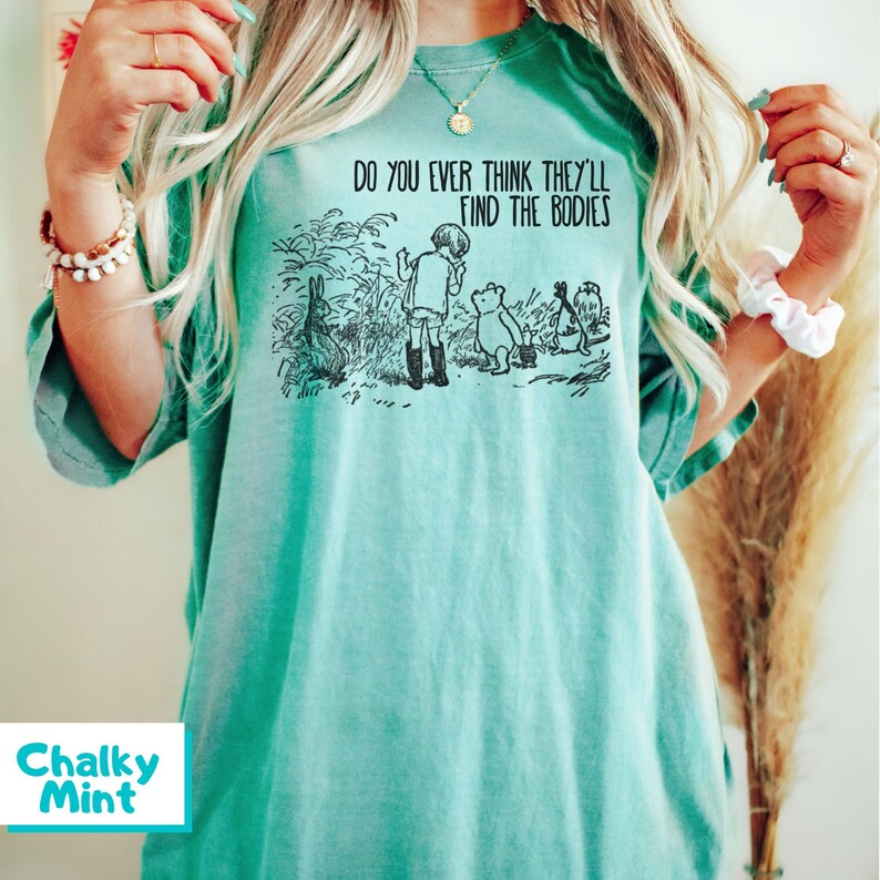 May include: A teal blue t-shirt featuring a black and white illustration of Winnie the Pooh and his friends. The text on the shirt reads "Do you ever think they'll find the bodies?"