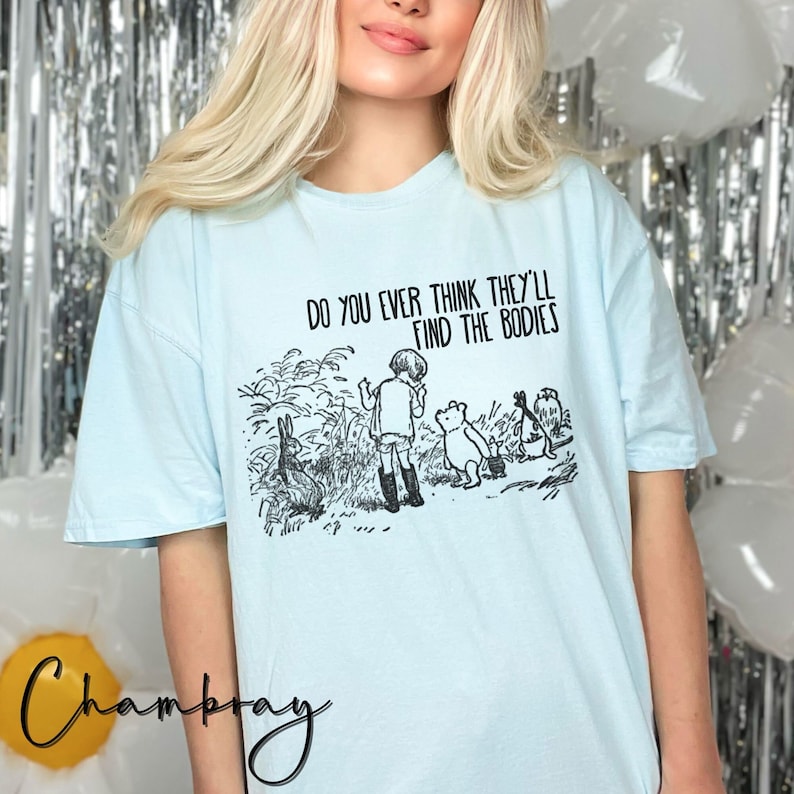 May include: A light blue t-shirt featuring a black and white illustration of Winnie the Pooh and his friends. The text "Do you ever think they'll find the bodies" is printed on the shirt.