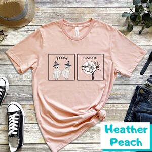 K&ouml;nnte beinhalten: Ein pinkfarbenes T-Shirt mit einem Aufdruck von zwei Geistern, die sich die H&auml;nde halten, vor dem Text "spooky" und einem Baum mit Flederm&auml;usen vor dem Text "season". Das Shirt liegt auf einer Holzoberfl&auml;che mit einer Jeans und einem Hut.
