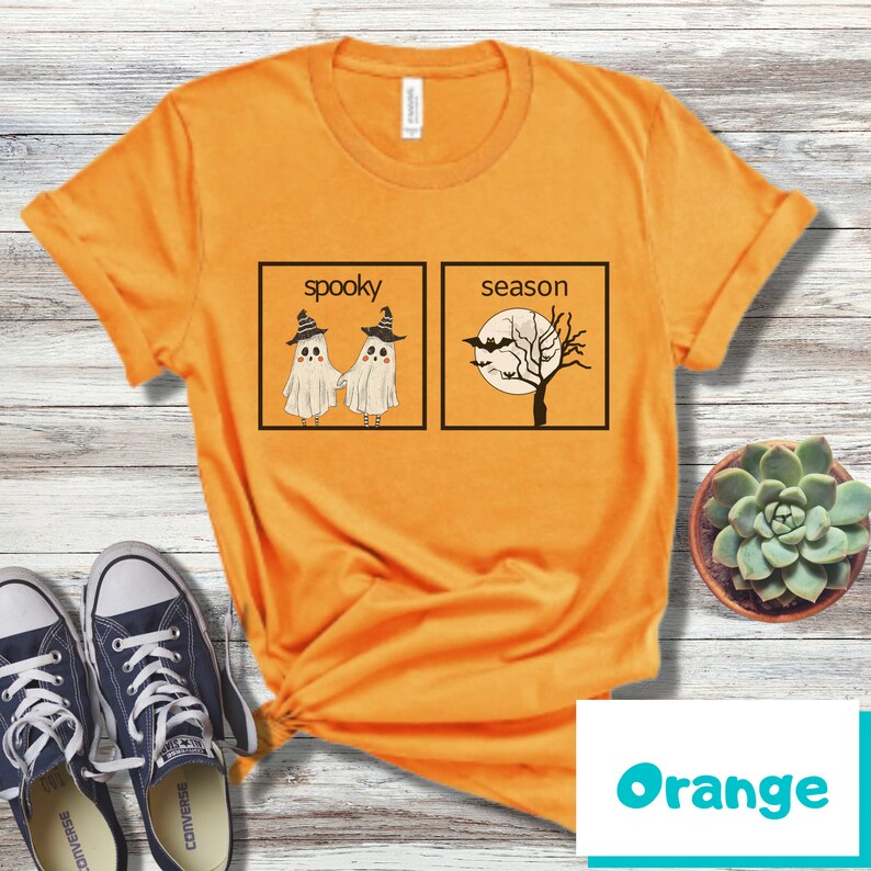 K&ouml;nnte beinhalten: Orangefarbenes T-Shirt mit einem Aufdruck von zwei Geistern, die Hexenh&uuml;te tragen, und dem Text "spooky" sowie einem Baum mit Flederm&auml;usen und dem Text "season" in schwarzen K&auml;stchen.
