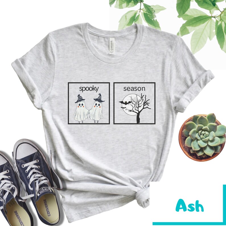 K&ouml;nnte beinhalten: Ein hellgraues T-Shirt mit einem grafischen Design, das zwei Geister zeigt, die sich in einer Box mit der Aufschrift "spooky" die H&auml;nde halten, und einen Baum mit Flederm&auml;usen in einer Box mit der Aufschrift "season".