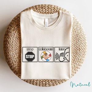 May include: A white t-shirt with a graphic design featuring three squares. The first square says "stop" with a red stop sign inside. The second square says "collaborate" with a graphic of hands of different skin tones holding each other. The third square says "listen" with a graphic of a hand over an ear.