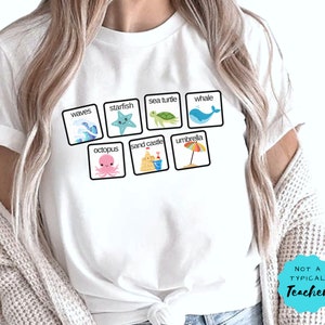 K&ouml;nnte beinhalten: Wei&szlig;es T-Shirt mit einer Grafik aus bunten Cartoon-Bildern von Meeresmotiven, darunter Wellen, Seesterne, Meeresschildkr&ouml;ten, Wale, Kraken, Sandburgen und Regenschirme. Der Text "NOT A TYPICAL TEACHER" ist unten rechts auf dem Shirt aufgedruckt.