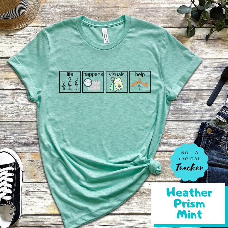 May include: A turquoise t-shirt with a graphic of four squares. Each square has a simple illustration and text. The text reads "life", "happens", "visuals", and "help". The t-shirt has a white tag with the text "Canvas" on the collar. The t-shirt is knotted and laid on a wooden surface. A white circle with the text "Not a Typical Teacher" is in the lower right corner. The text "Heather Prism Mint" is below the circle.