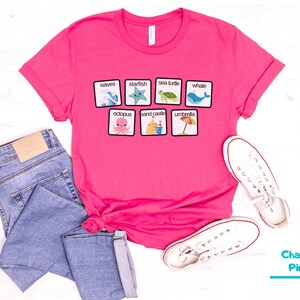 K&ouml;nnte beinhalten: Pinkes T-Shirt mit einem Grafikdruck von einem Satz Strand-Bildkarten. Die Karten zeigen Bilder von Wellen, Seesternen, Meeresschildkr&ouml;ten, Walen, Kraken, Sandburgen und Sonnenschirmen. Der Text auf den Karten lautet: "Wellen", "Seestern", "Meeresschildkr&ouml;te", "Wal", "Kraken", "Sandburg" und "Sonnenschirm".
