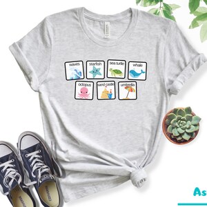 K&ouml;nnte beinhalten: Ein hellgraues T-Shirt mit einer Grafik von Strand-Icons in Quadraten. Die Icons beinhalten Wellen, einen Seestern, eine Meeresschildkr&ouml;te, einen Wal, einen Oktopus, eine Sandburg und einen Regenschirm. Der Text "waves", "starfish", "sea turtle", "whale", "octopus", "sand castle", und "umbrella" ist unter jedem Icon gedruckt.