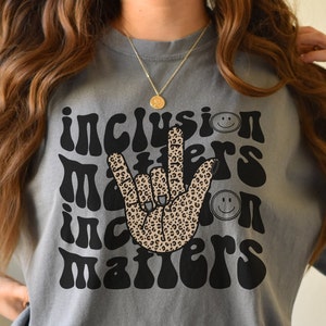 May include: Gray t-shirt with a black and white graphic design of a hand making the 'I love you' sign in American Sign Language. The hand is covered in a leopard print pattern. The text 'inclusion matters' is written in a retro font around the hand.