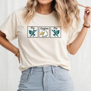May include: A cream-colored short-sleeve t-shirt featuring a graphic print of three square boxes. The outer boxes contain a teal superhero figure with the text Fly, while the center box displays an eagle head with the text Eagles.