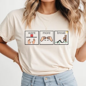 Puede incluir: Una camiseta de manga corta color crema con tres paneles gráficos rectangulares. Los paneles contienen el texto "want", "more" y "break" acompañados de ilustraciones sencillas de manos, un bloque rojo y una figura de palo descansando junto a un árbol.