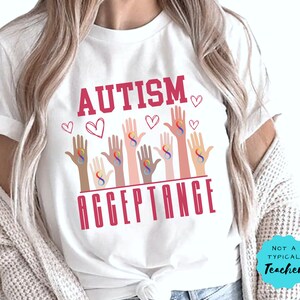 May include: White t-shirt with the text "AUTISM ACCEPTANCE" in pink lettering. The design features multiple hands with rainbow infinity symbols, representing autism awareness.