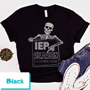 May include: Black t-shirt featuring a skeleton holding a sign that reads "IEP SEASON" with the text "I'll sleep when I'm dead." The design is in gray and white. The shirt is paired with blue jeans and white sneakers.