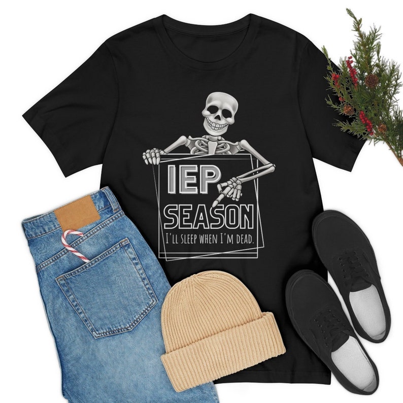 May include: Black t-shirt with a skeleton graphic holding a sign that reads "IEP SEASON" with the text "I'll sleep when I'm dead." Also pictured are blue jeans, a tan beanie, and black shoes.
