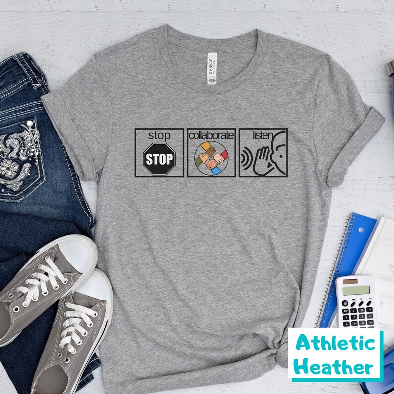 May include: Gray t-shirt with the words "stop", "collaborate", and "listen" in black text. Each word is in a white box with a symbol inside. The symbol for "stop" is a red stop sign. The symbol for "collaborate" is a circle with five hands of different skin tones holding each other. The symbol for "listen" is a hand cupped over an ear. The text "Athletic Heather" is in a blue box in the bottom right corner of the image.