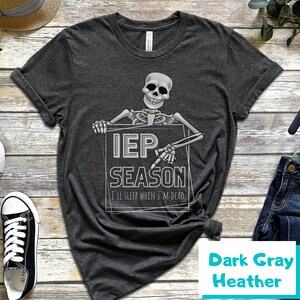 May include: Dark gray heather t-shirt with a skeleton graphic holding a sign that reads "IEP SEASON" and "I'll sleep when I'm dead". The shirt is a dark gray color and has a classic crew neck.