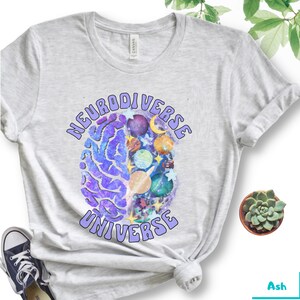 Może przedstawiać: Szara koszulka z nadrukiem fioletowego m&oacute;zgu i tekstem "Neurodiverse Universe" w fioletowym, retro stylu.