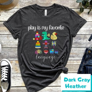 May include: Dark gray heather t-shirt with the text "Play is my favorite language" and a graphic of colorful toys, including a toy dinosaur, a toy robot, a toy train, and a toy duck.