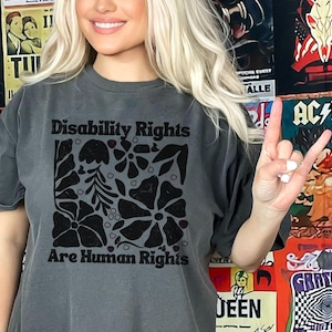 May include: A gray t-shirt with the text "Disability Rights Are Human Rights" in black. The shirt features a black floral design. The person is giving a rock and roll hand gesture.