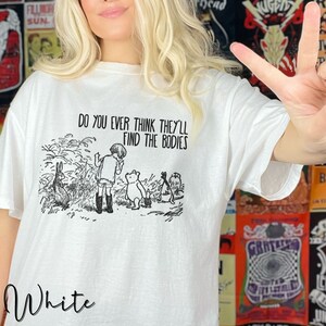 May include: White t-shirt featuring a black and white illustration of Winnie the Pooh and his friends. The text "Do you ever think they'll find the bodies" is printed on the shirt.