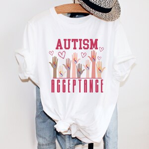 May include: White t-shirt with the words "AUTISM ACCEPTANCE" in red lettering. The design features multiple hands of different skin tones, each with a rainbow infinity symbol.