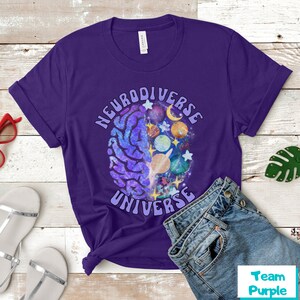 Może przedstawiać: Fioletowy T-shirt z grafiką m&oacute;zgu z planetami i gwiazdami w środku. Wok&oacute;ł m&oacute;zgu napisano "Neurodiverse Universe".