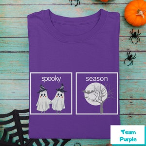 K&ouml;nnte beinhalten: Lilafarbenes T-Shirt mit einer wei&szlig;en Grafik von zwei Geistern mit Hexenh&uuml;ten und dem Text "spooky" und einem Baum mit einer Fledermaus und dem Text "season".