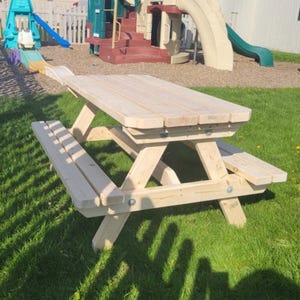 May include: A wooden picnic table with attached benches. The table and benches are made of light-colored wood and are set on a grassy lawn. The table is rectangular with rounded corners. The benches are attached to the table.