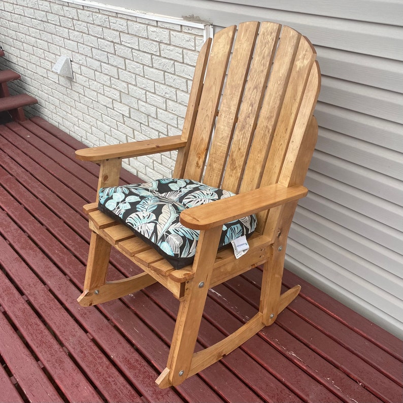 May include: A wooden rocking chair with a light brown finish and a blue and white floral cushion. The chair is on a wooden deck.